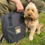 Dog sitting next to a black backpack with 'Made for Adventure' text on grass