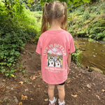 Child wearing a pink t-shirt with a nature-themed design, standing in a forested area.