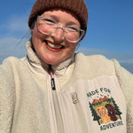 Person wearing a brown knit hat with a pom-pom and a beige fleece jacket with a dog graphic and text, outdoors against a blue sky.