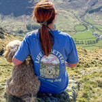 Person wearing a blue t-shirt with a dog graphic and text, sitting on a mountain with a scenic view.