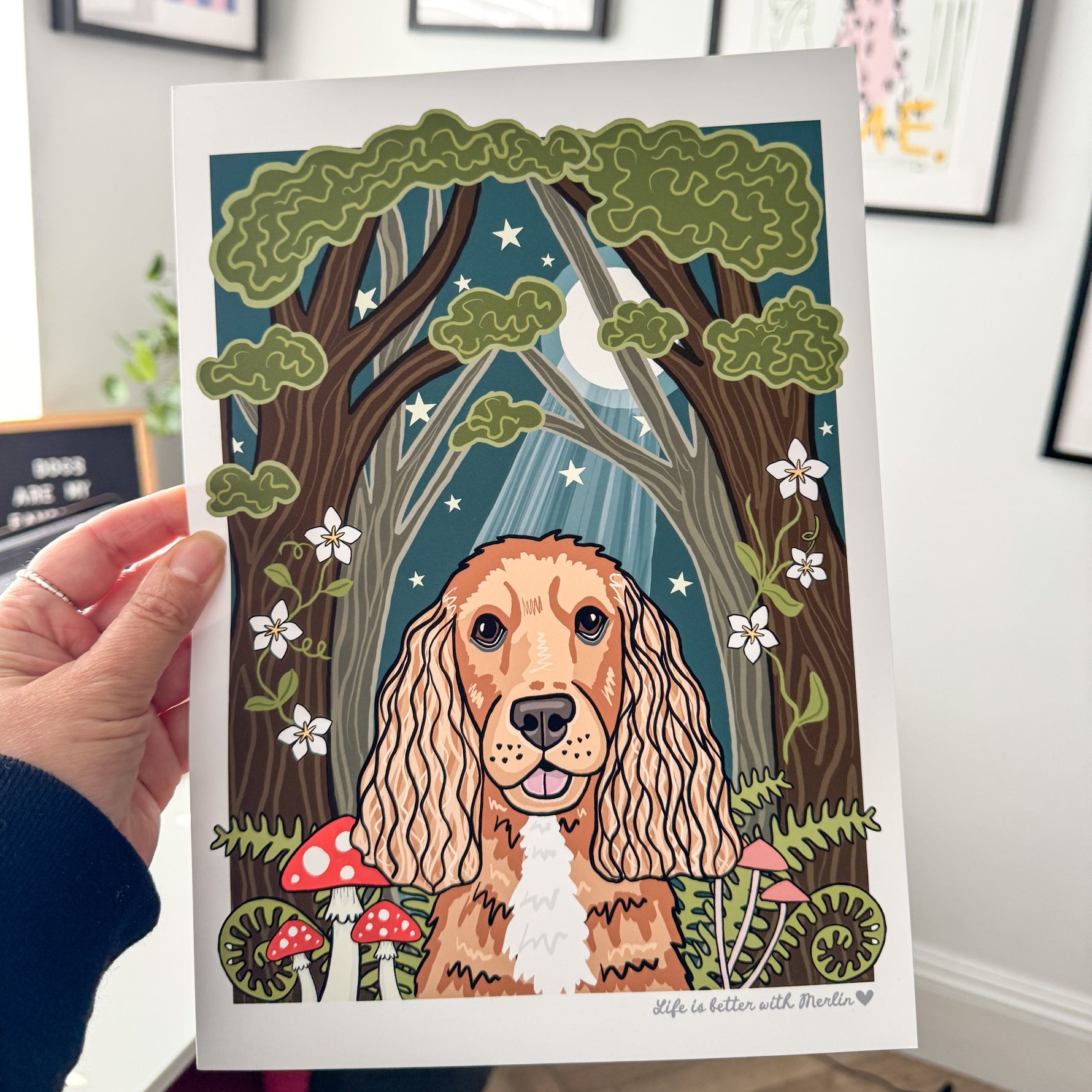 Person holding a print of a dog in a forest illustration.