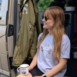 Woman sitting in a vehicle holding a mug, wearing a t-shirt with graphics and sunglasses.