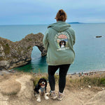 Person wearing a green jacket with 'Life is Better' text and dog standing on a cliff overlooking the sea.