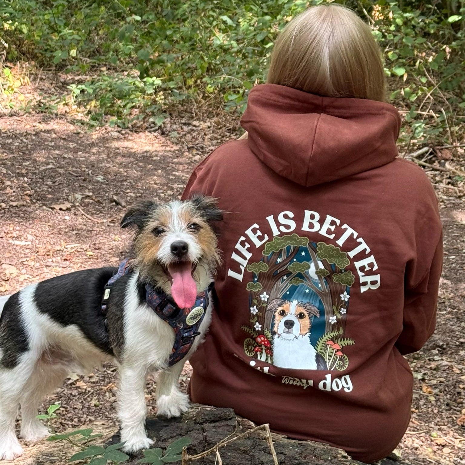 Person wearing a hoodie with a dog graphic and 'Life is Better' text, standing in a forest with a dog.