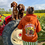A person wearing a rust orange hoodie with a dog-themed graphic that reads "life is better with my dogs" stands in a sunflower field next to a red tractor, with two brown dogs sitting on the tractor tire.