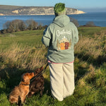 Person wearing a hoodie with 'Outside' text and a dog walking on a grassy field by the sea.