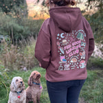A woman viewed from behind wearing a mauve-brown hoodie with a colorful 'Feel Good Walking Club' graphic on the back, standing outdoors with two dogs on a grassy field at sunset.
