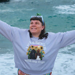 A woman stands on a beach with her arms outstretched, wearing a lavender sweatshirt with a graphic print that reads 'life is Better with my dogs' and a colorful patterned bikini. The ocean and rocks are visible in the background.