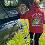 Person in a 'Made for Adventure' hoodie with a dog on a vehicle roof rack in a grassy area.