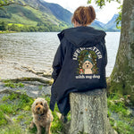 Person wearing a black jacket with a dog graphic and text by a lake with mountains in the background