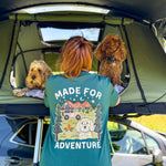 Person with 'Made for Adventure' shirt standing next to a car with two dogs inside a tent.