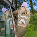 Person wearing a green jacket with 'Outside' text and nature-themed graphics, standing next to a vehicle.