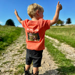 Child in an orange shirt with a graphic design, standing on a dirt path with arms raised against a blue sky.