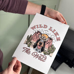 Person holding a printed sheet with a dog illustration and 'Wild Like the West' text.
