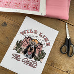 Print with 'Wild Like The West' design on a wooden surface with scissors and fabric.