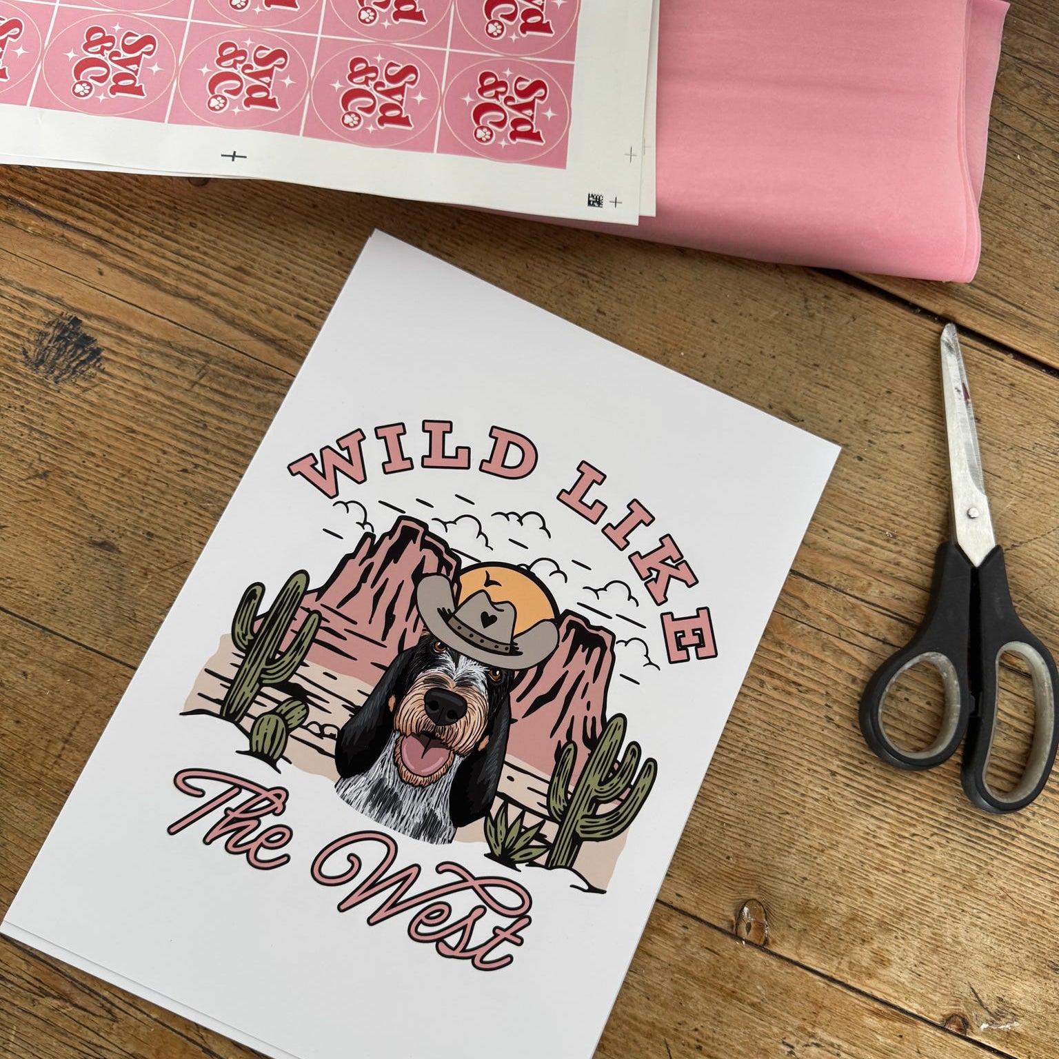 Print with 'Wild Like The West' design on a wooden surface with scissors and fabric.