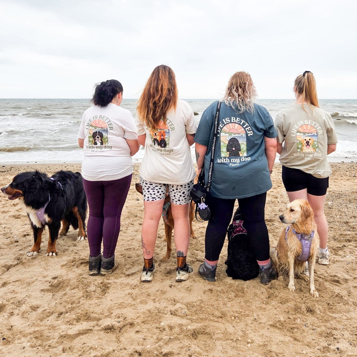 Four people and two dogs on a beach wearing shirts with visible text and graphics.