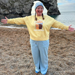 Person wearing a yellow sweatshirt with a graphic design, standing on a beach with a large rock formation and ocean in the background.