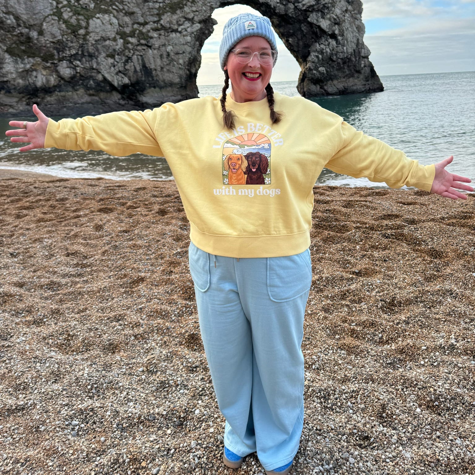 Person wearing a yellow sweatshirt with a graphic design, standing on a beach with a large rock formation and ocean in the background.