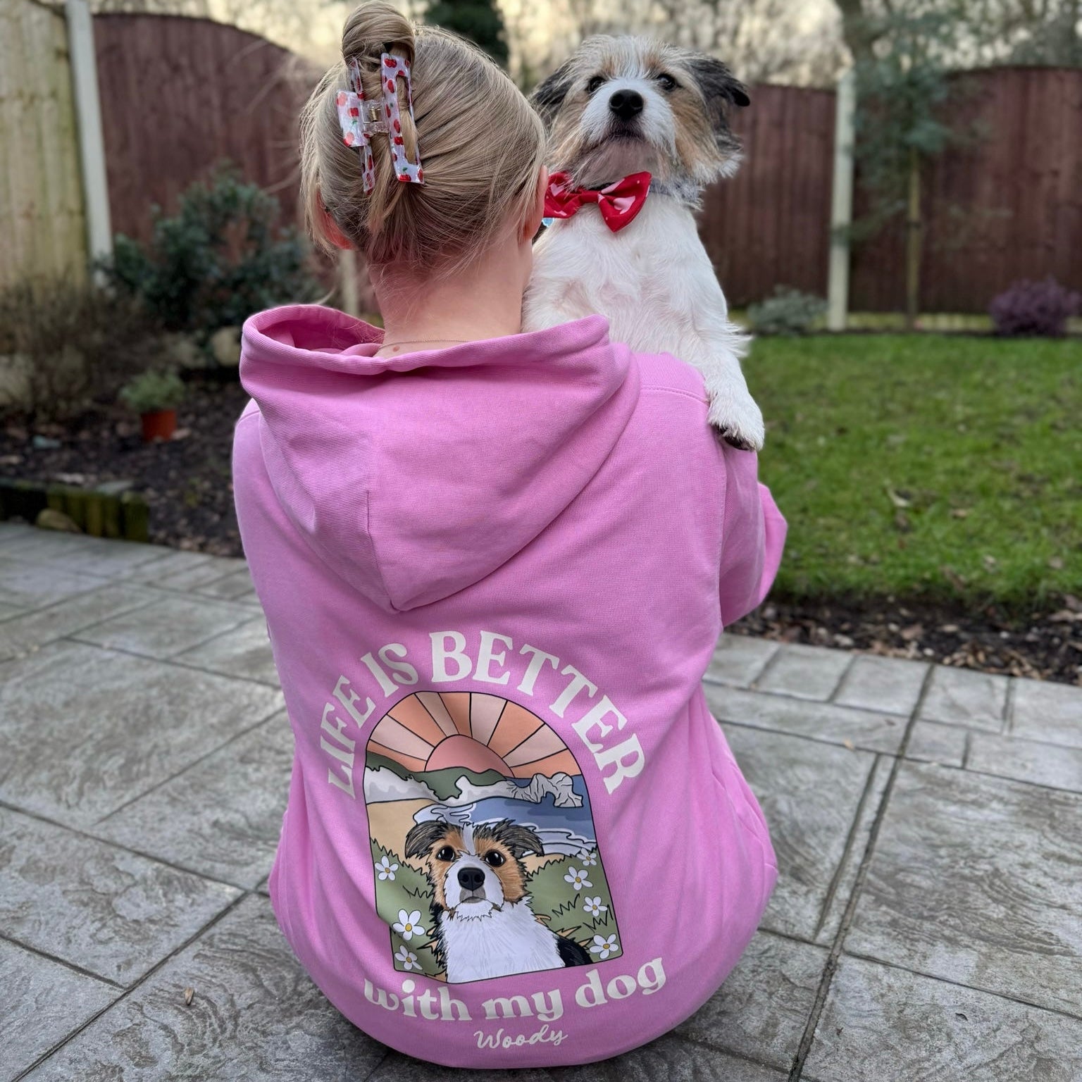 Person wearing a pink hoodie with a dog graphic and text, holding a small dog in an outdoor setting.