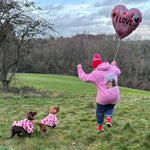 Child in pink jacket and hat holding a heart-shaped balloon with dogs in polka dot coats on a grassy field.