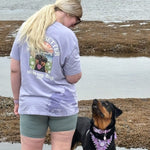 Woman wearing a t-shirt with a dog graphic and text, standing next to a dog by a body of water.