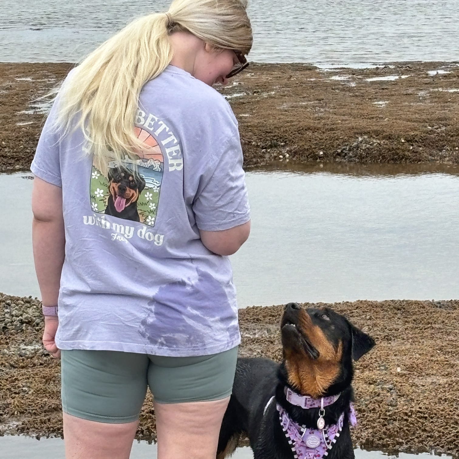 Woman wearing a t-shirt with a dog graphic and text, standing next to a dog by a body of water.