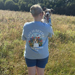 Person holding a dog in a field wearing a shirt with a dog graphic and text.