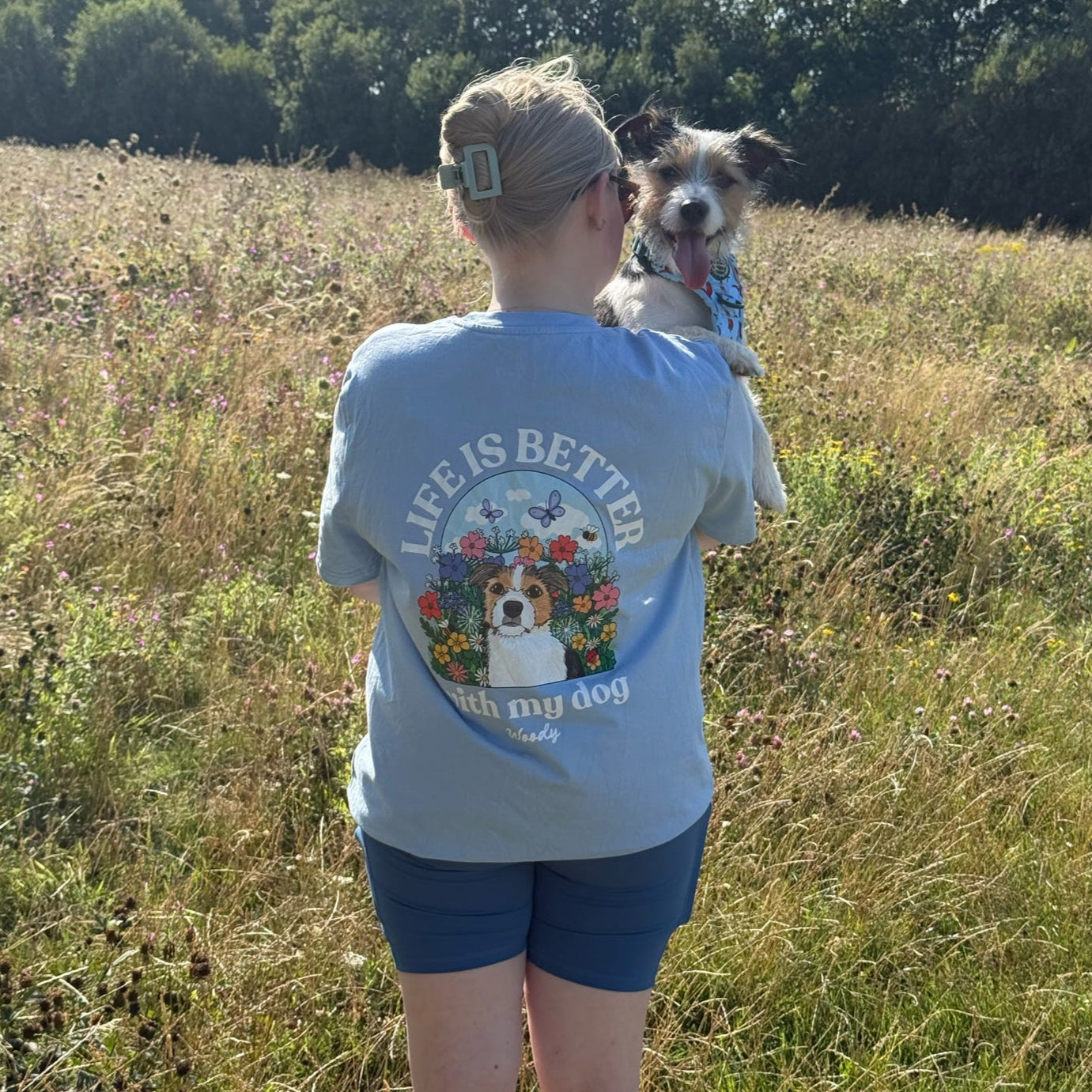 Person holding a dog in a field wearing a shirt with a dog graphic and text.