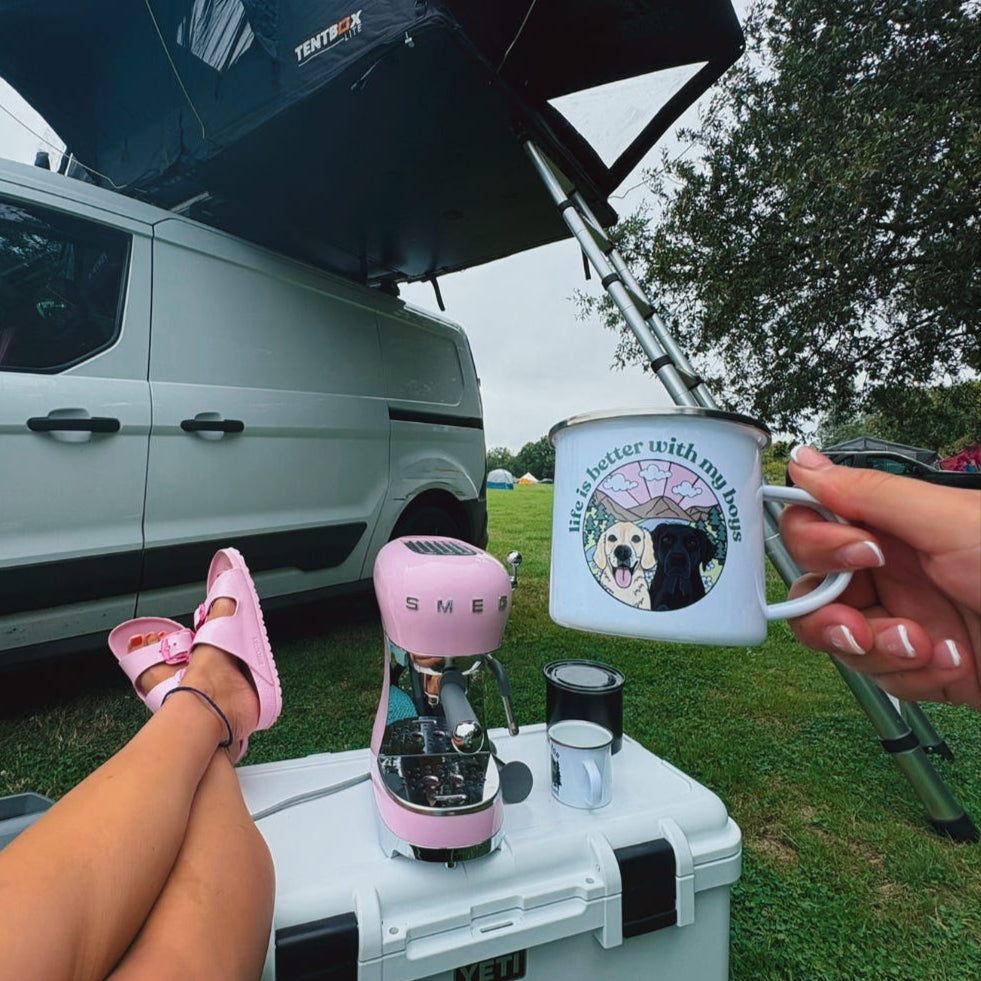 Person sitting on a cooler with a pink water bottle and a mug, next to a van with an open roof tent.