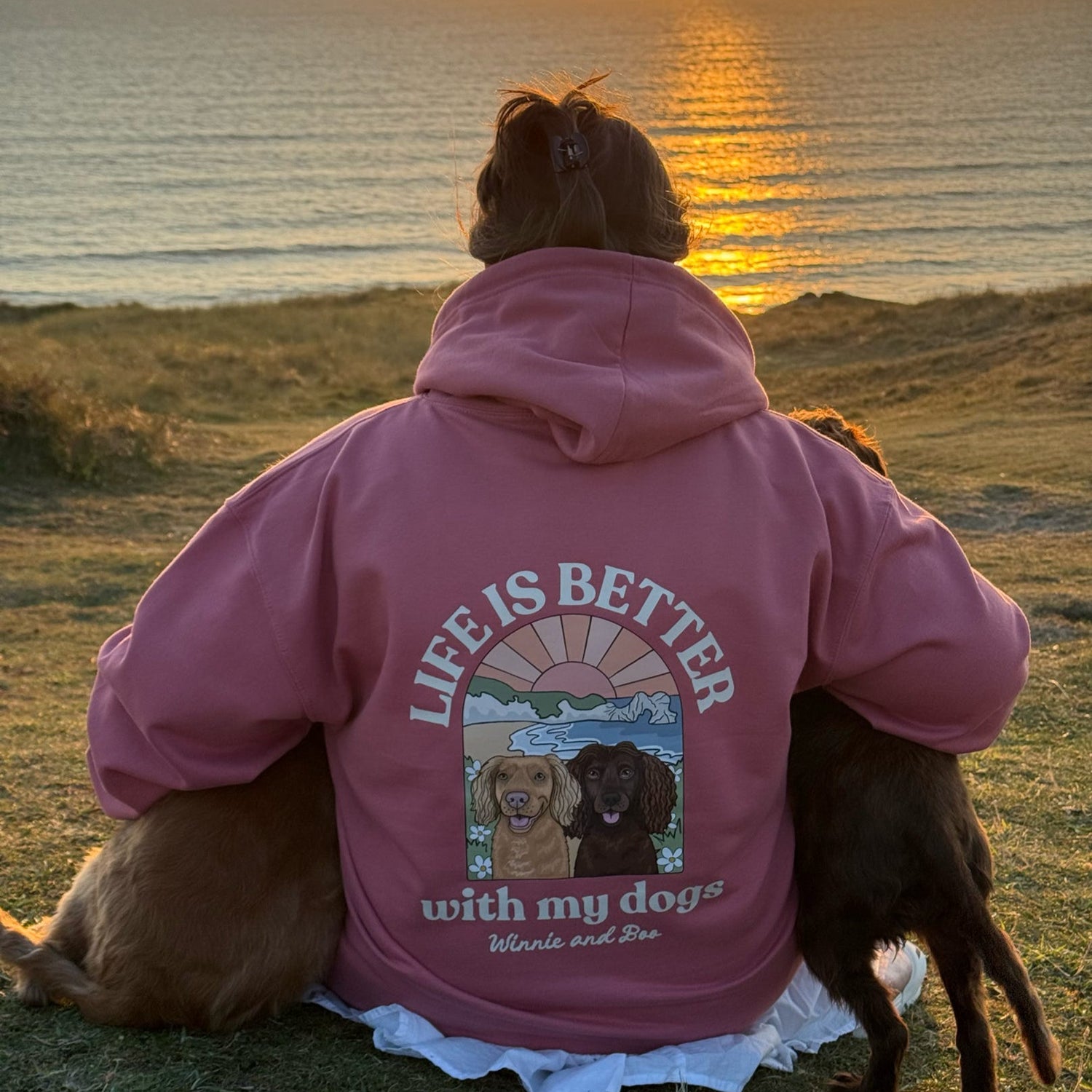 Person wearing a pink hoodie with a dog-themed design sitting by the ocean at sunset.