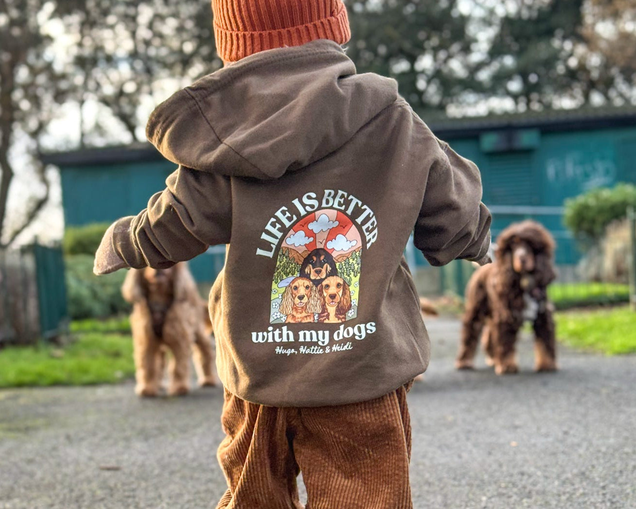 Child wearing a brown hoodie with a dog-themed design and text, walking outdoors with dogs in the background.