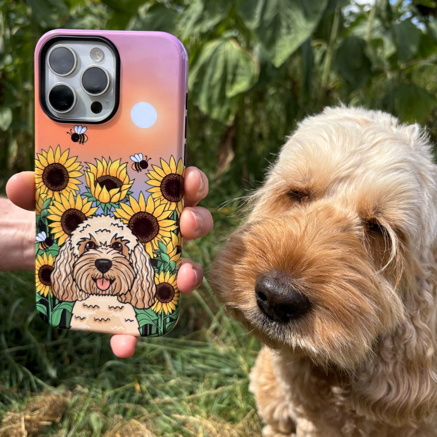 A hand holds a phone case featuring a dog illustration and sunflowers, with a real dog standing outdoors in a garden background.