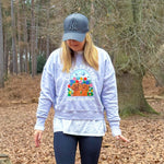 A woman walking in a wooded area covered with brown leaves, wearing a light purple graphic sweatshirt, a gray baseball cap, dark leggings, and yellow sneakers.