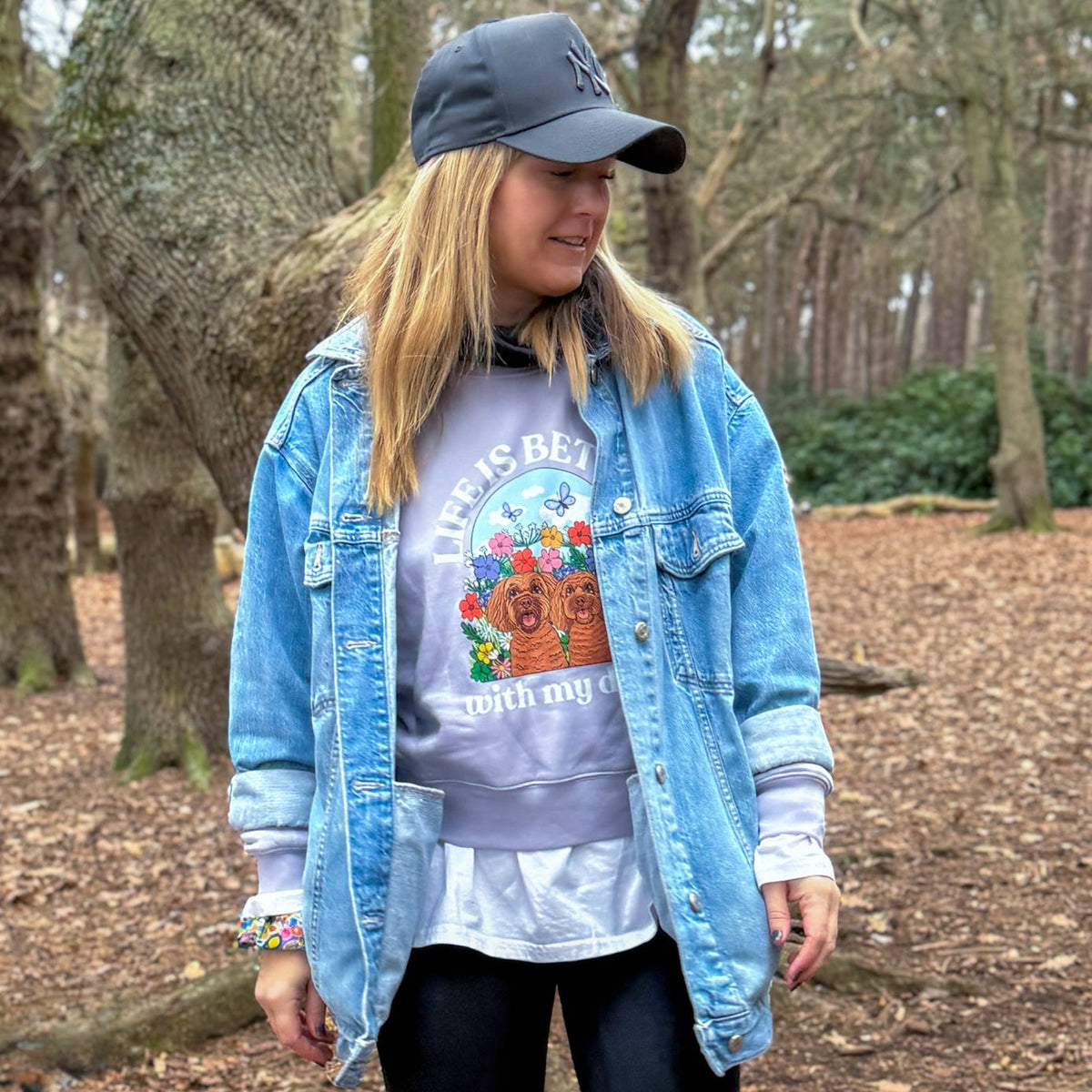 A woman standing in a wooded outdoor setting wearing a light blue denim jacket over a graphic sweatshirt, a dark baseball cap, black leggings, and white socks.