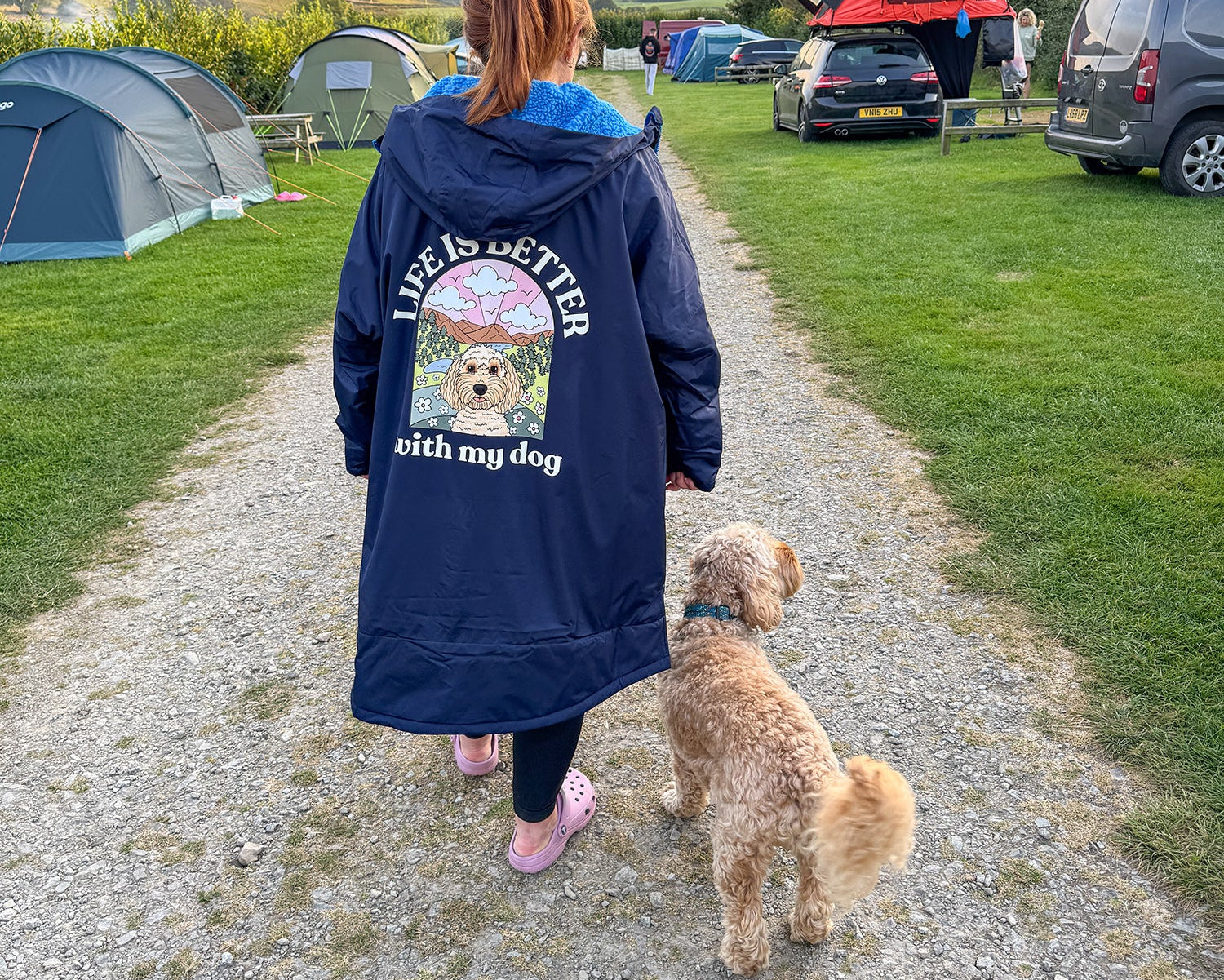 Person walking a dog on a path with tents and vehicles in the background at a camping site.