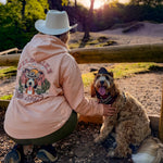 Person wearing a pink hoodie with a dog sitting on the ground outdoors during sunset.