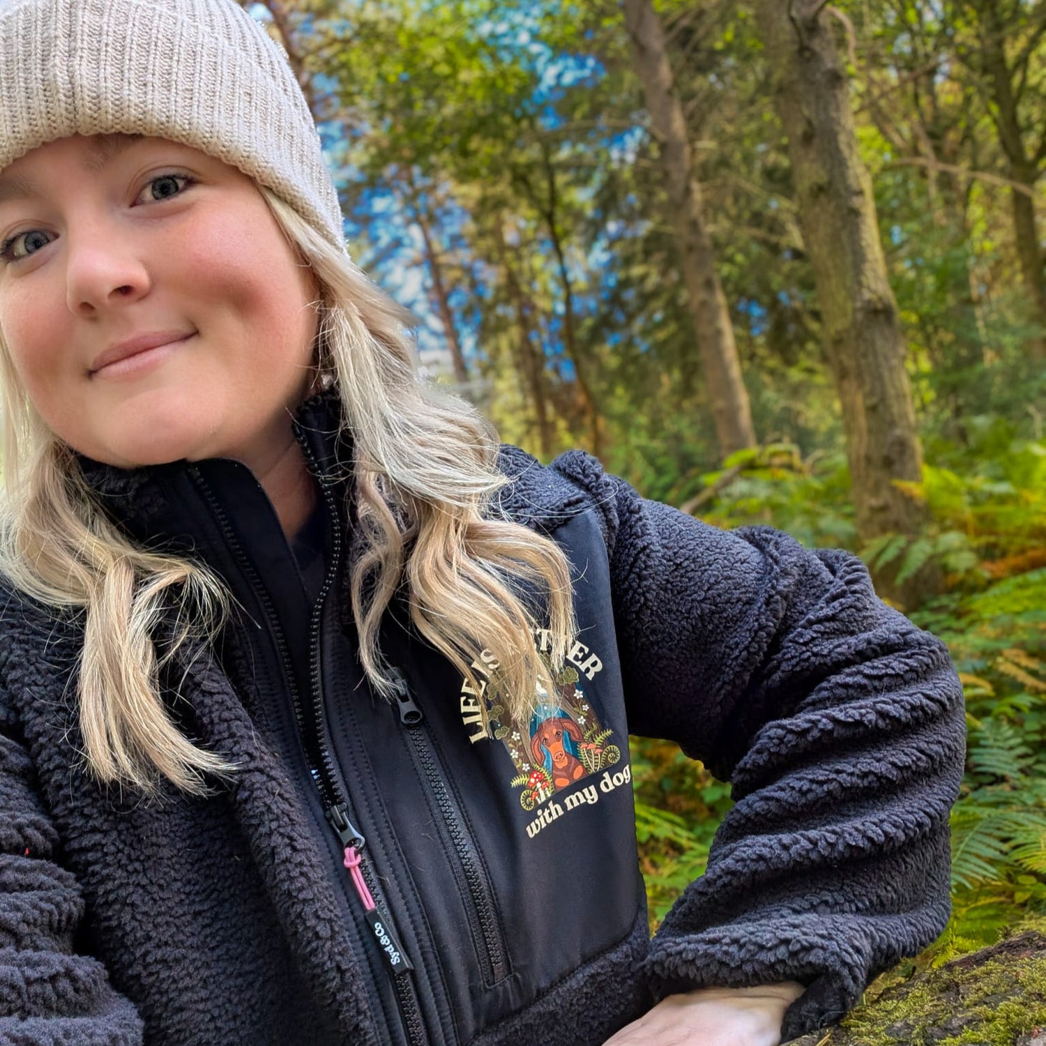 Woman in a forest setting wearing a black jacket and beige beanie.