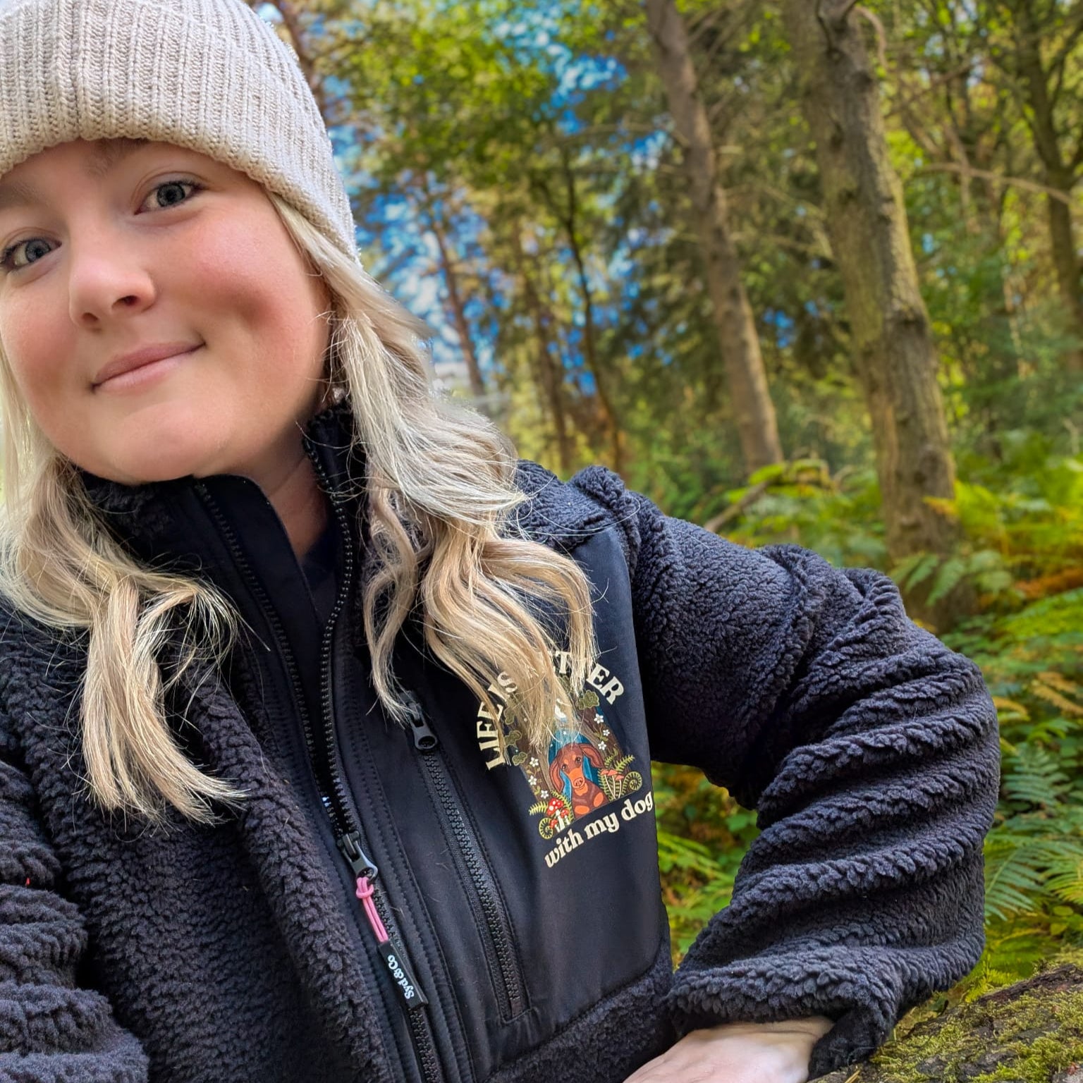 Woman in a forest setting wearing a black jacket and beige beanie.