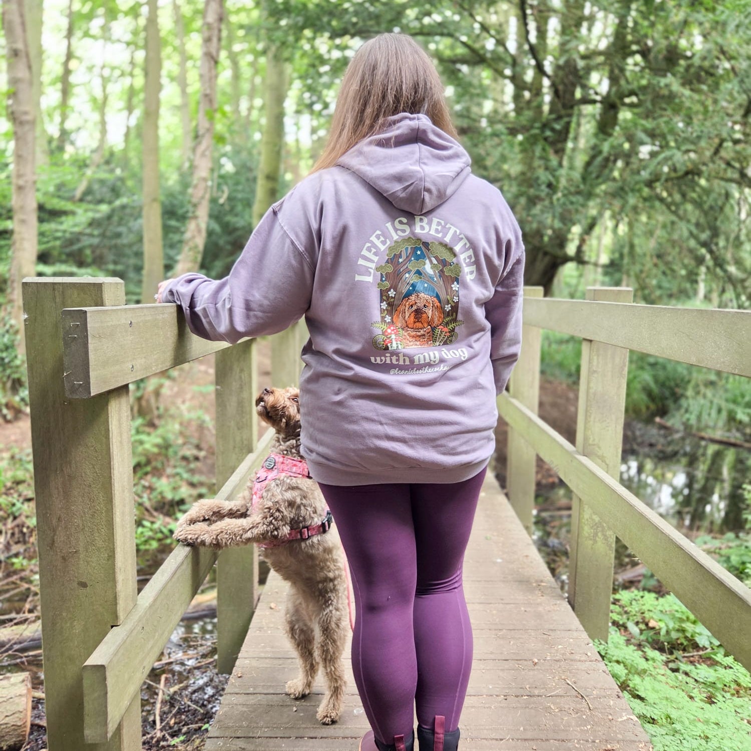 Person wearing a hoodie with a dog design, standing on a wooden bridge in a forest.
