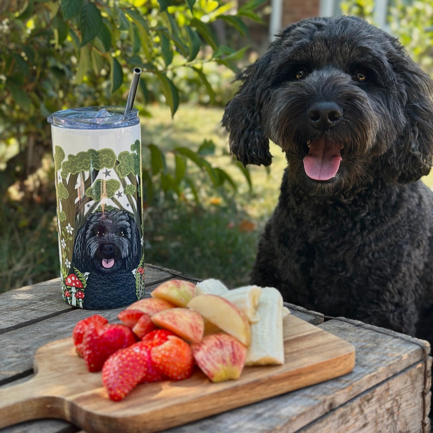 Dog sitting next to a tumbler with a dog design and a wooden board with fruits outdoors.