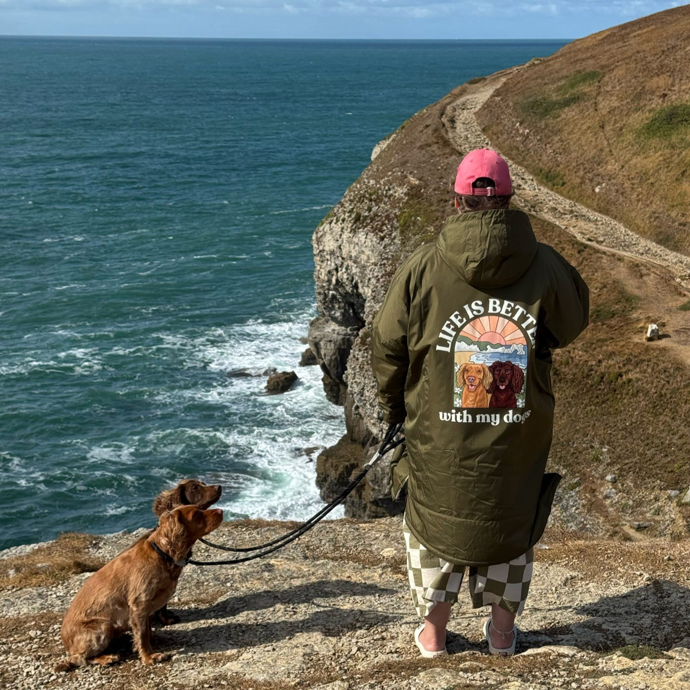 Person wearing a green jacket with a dog-themed design, standing on a cliff overlooking the ocean.