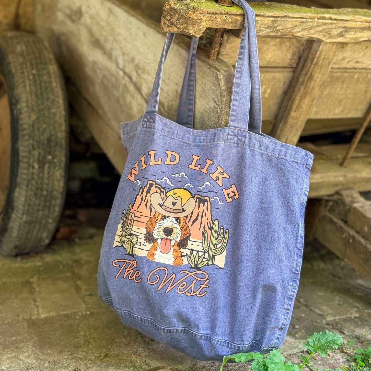 Blue tote bag with 'Wild Like The West' design hanging on a wooden cart.