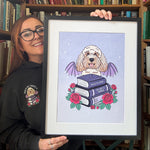 A woman wearing glasses and a black hoodie holds up a framed art print featuring a fluffy white dog with purple wings perched on a stack of books, surrounded by red roses, set against a background of bookshelves.