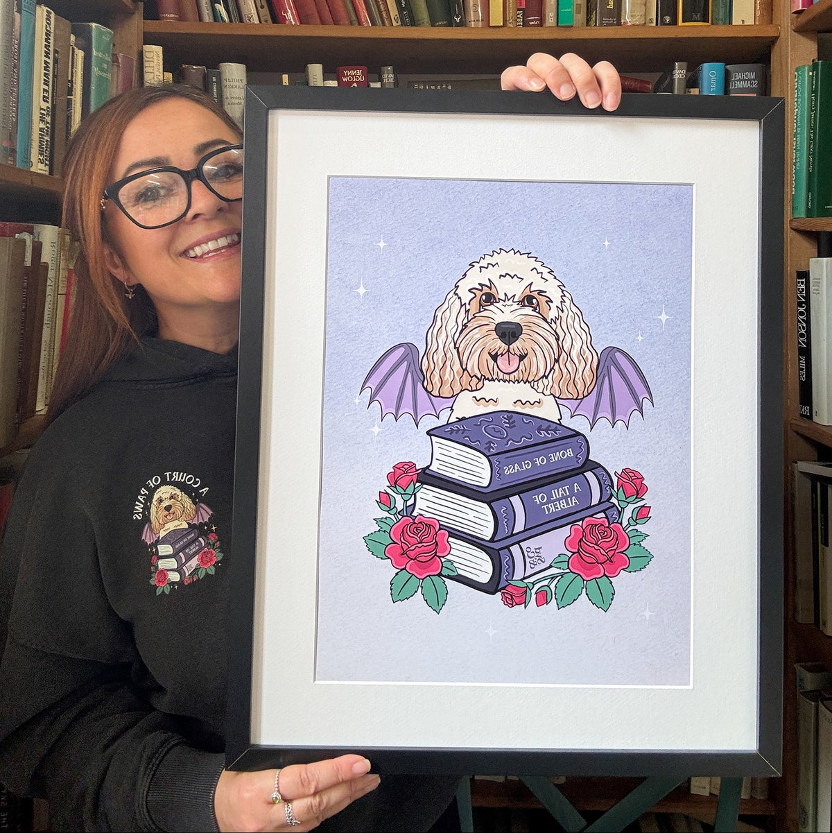A woman wearing glasses and a black hoodie holds up a framed art print featuring a fluffy white dog with purple wings perched on a stack of books, surrounded by red roses, set against a background of bookshelves.