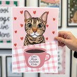 Card with a cat illustration and 'Cat Mum Coffee Club' text held by a hand in front of framed pictures.