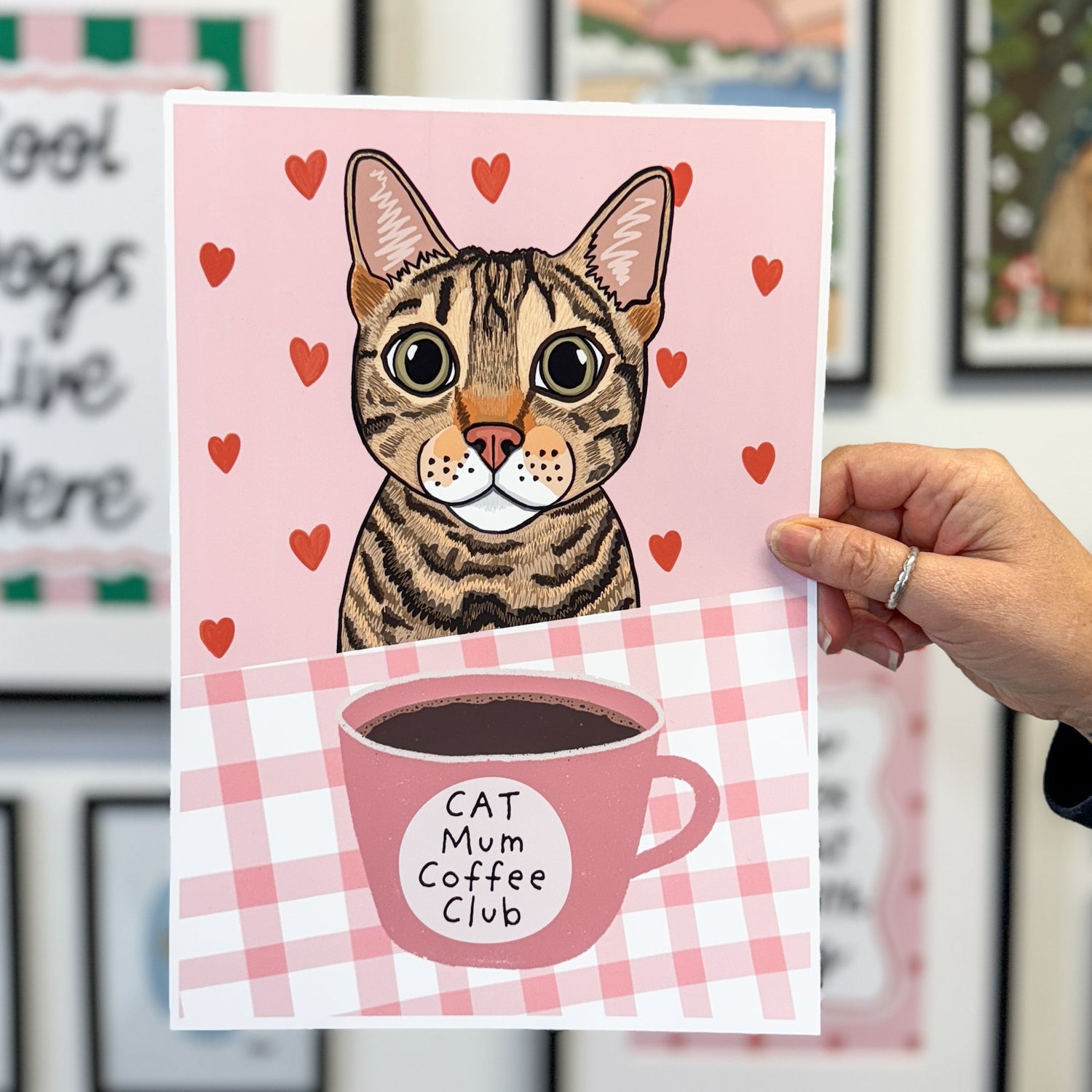 Card with a cat illustration and 'Cat Mum Coffee Club' text held by a hand in front of framed pictures.
