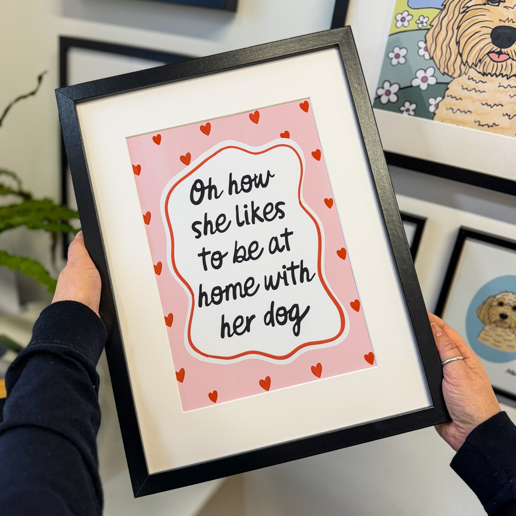 Person holding a framed print with a quote about being at home with a dog, surrounded by other framed artwork.