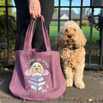 A person holding a dusty mauve canvas tote bag next to a small fluffy cream-colored dog, set outdoors in front of a black metal gate.