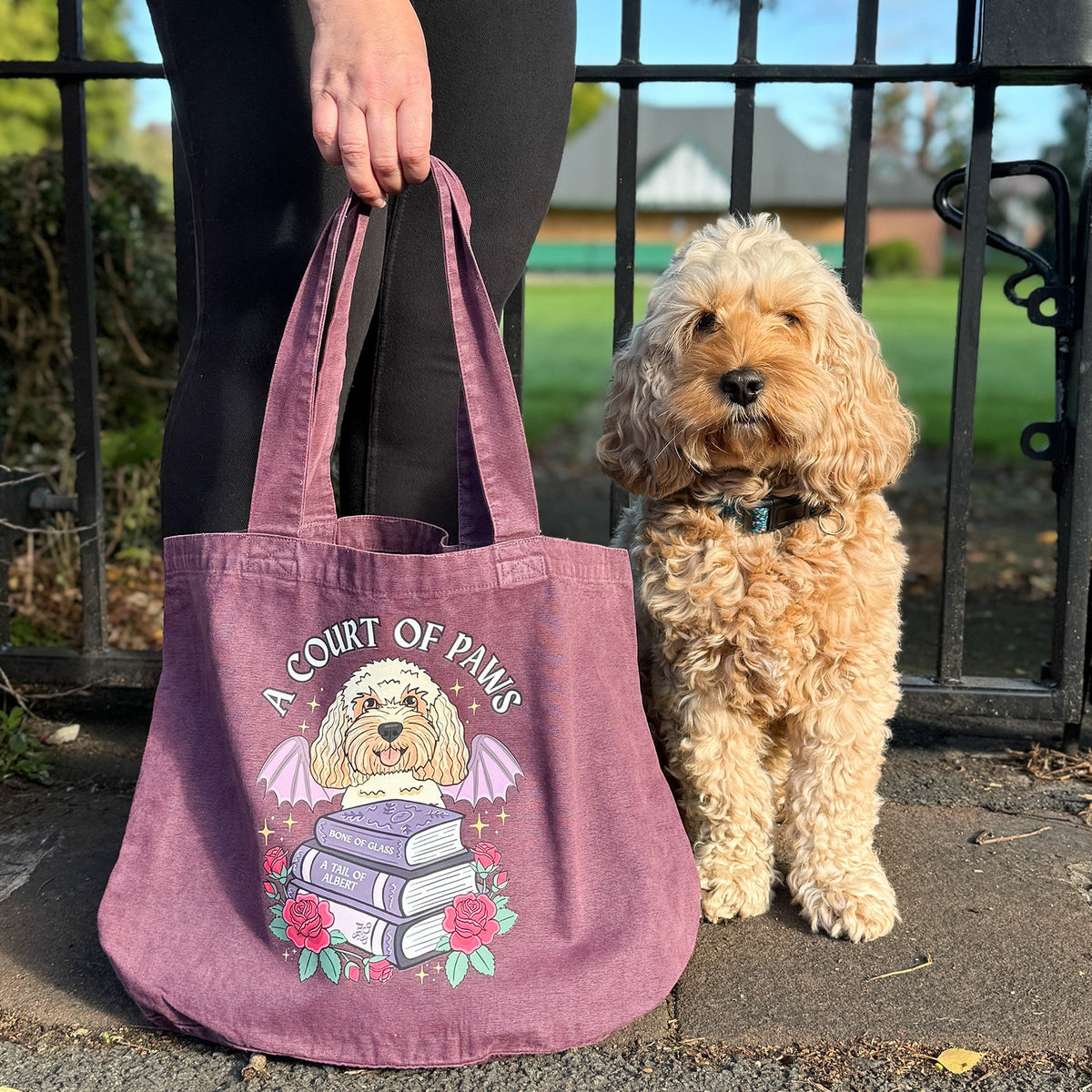 A person holding a dusty mauve canvas tote bag next to a small fluffy cream-colored dog, set outdoors in front of a black metal gate.
